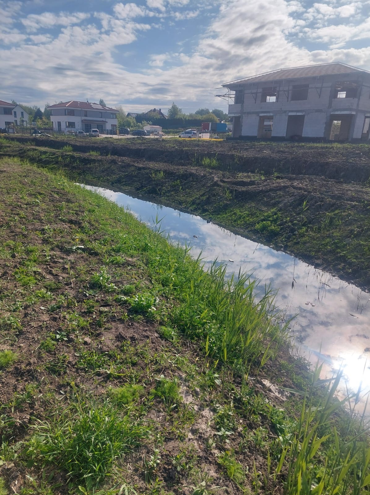 Melioracja rowu w Szczecinie. Woda w rowie odbija błękitne niebo z chmurami. W tle widać budynki w trakcie budowy i inne zabudowania.