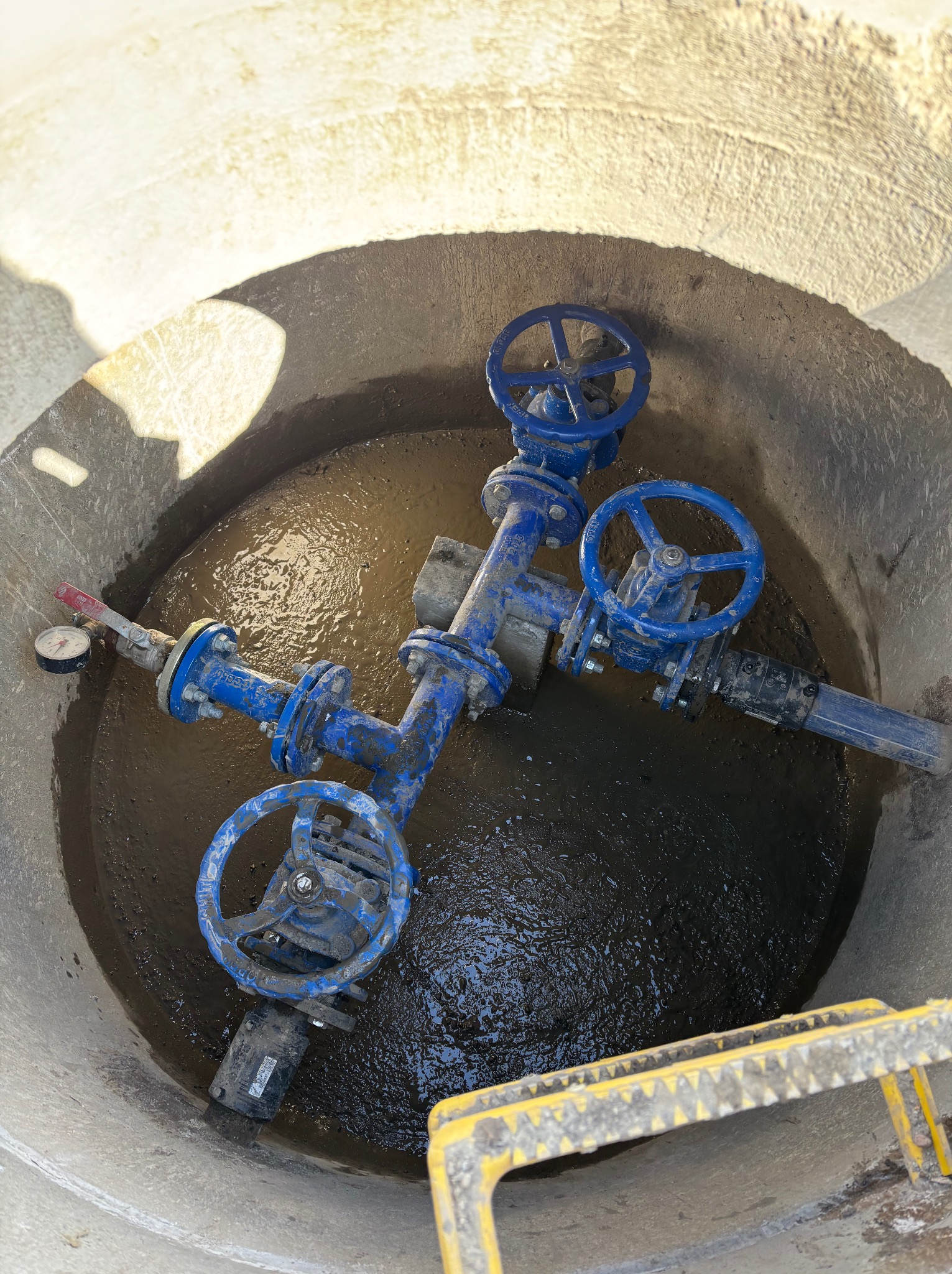 Studzienka rewizyjna z niebieską armaturą wodociągową i manometrem, częściowo zalana wodą. Widoczny fragment drabiny ze szczeblami.