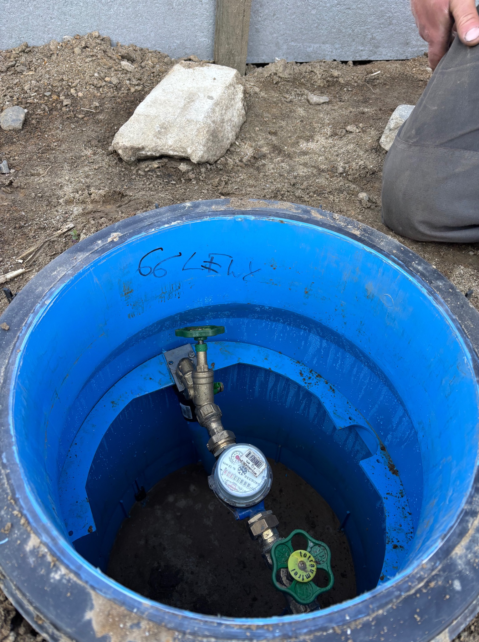 Niebieski właz studzienki z wodomierzem i zaworami, widoczne elementy instalacji podziemnej. W tle ziemia i fragment betonowego ogrodzenia.