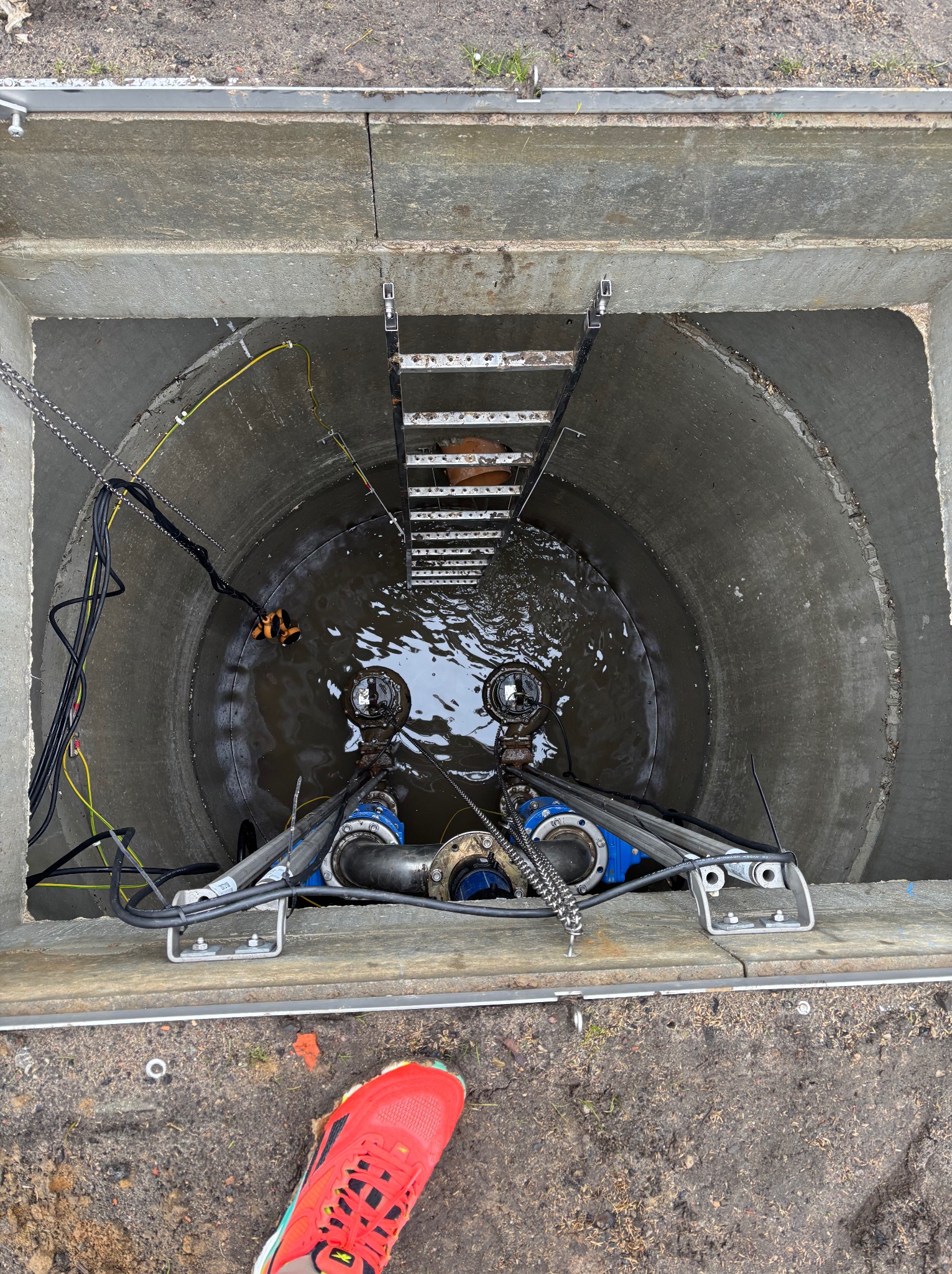 Betonowy zbiornik studzienny z zainstalowanymi pompami zanurzeniowymi i drabinką, widok z góry, kabel zasilający, czerwony but w rogu kadru.