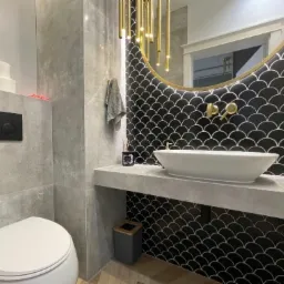 Nowoczesna łazienka z czarnymi płytkami w kształcie rybiej łuski, owalną białą umywalką nablatową, złotą armaturą i wiszącą lampą z podłużnych, złotych elementów, szara toaleta w tle.