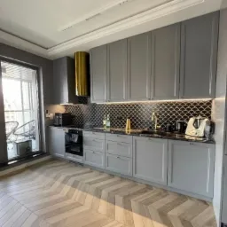 Nowoczesna kuchnia w odcieniach szarości z parkietem w jodełkę, czarną mozaiką w kształcie rybiej łuski nad blatem i złotym okapem, widok na balkon z krzesłami.