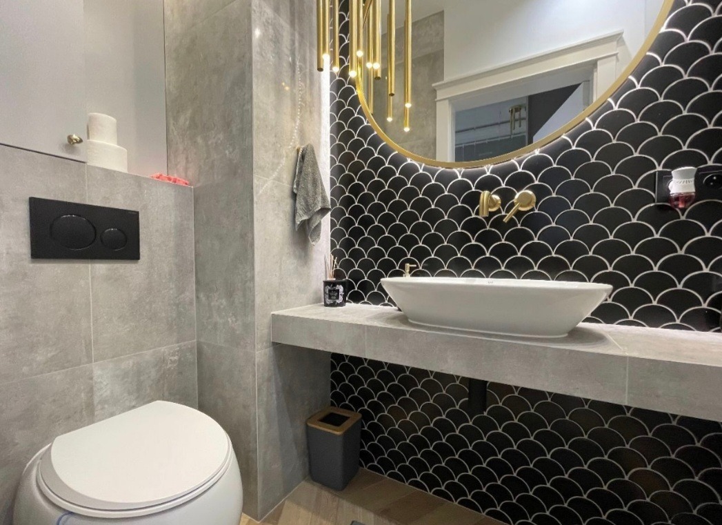 Nowoczesna łazienka z czarnymi płytkami w kształcie rybiej łuski, owalną białą umywalką nablatową, złotą armaturą i wiszącą lampą z podłużnych, złotych elementów, szara toaleta w tle.