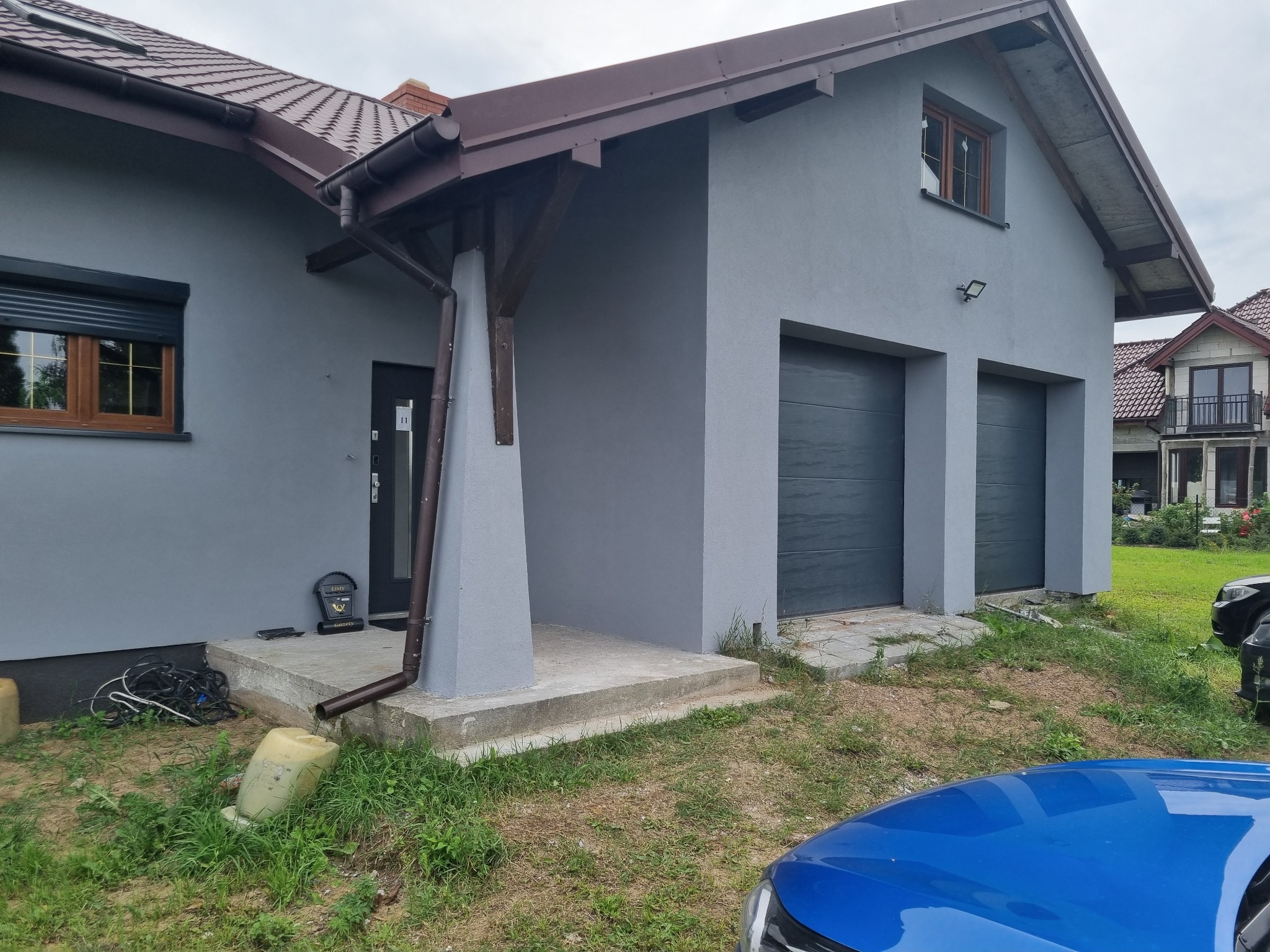 Dom jednorodzinny z szarą elewacją, brązowymi oknami i dwiema bramami garażowymi; widoczny fragment niebieskiego samochodu na pierwszym planie.