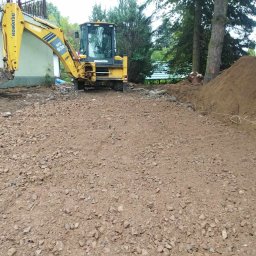 FHU Michał Sobesto - Żółta koparko-ładowarka Komatsu WB93R na placu budowy z przygotowanym terenem pod fundamenty, widoczny wykop i pryzma ziemi w tle, drzewa i budynek w tle.