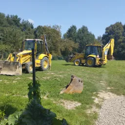 Dwa żółte traktory Komatsu WB97R Utility na trawiastym terenie z drzewami w tle, dodatkowo widoczna łyżka koparki na pierwszym planie.