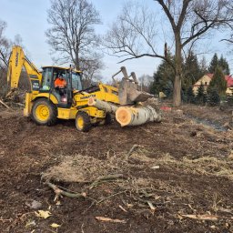 FHU Michał Sobesto - Żółta koparko-ładowarka Komatsu transportuje ścięty pień drzewa na błotnistym terenie po wycince. W tle widoczne drzewa i budynek mieszkalny.