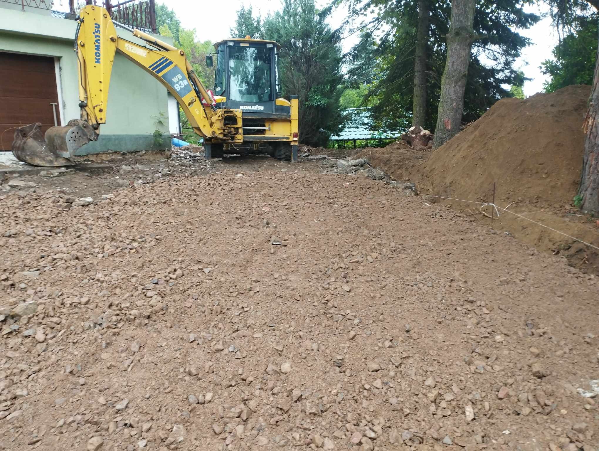 Żółta koparko-ładowarka Komatsu WB93R na placu budowy z przygotowanym terenem pod fundamenty, widoczny wykop i pryzma ziemi w tle, drzewa i budynek w tle.