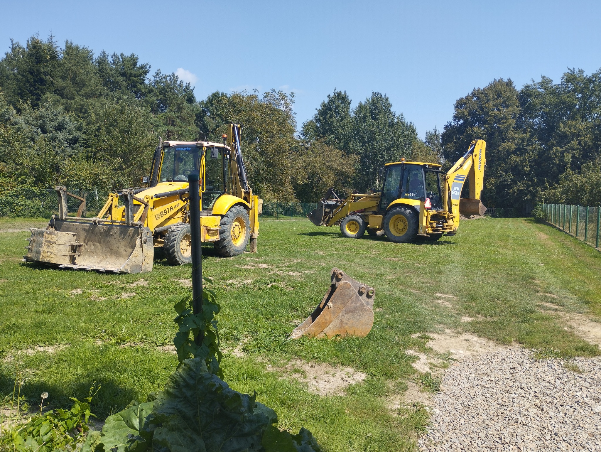 Dwa żółte traktory Komatsu WB97R Utility na trawiastym terenie z drzewami w tle, dodatkowo widoczna łyżka koparki na pierwszym planie.