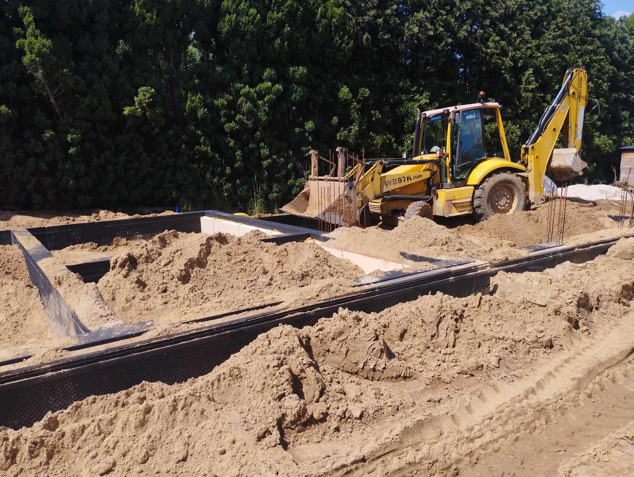 Żółta koparko-ładowarka WB97R zasypuje piaskiem szalunek fundamentowy z czarną izolacją, widoczne wystające pręty zbrojeniowe i zielone drzewa w tle.
