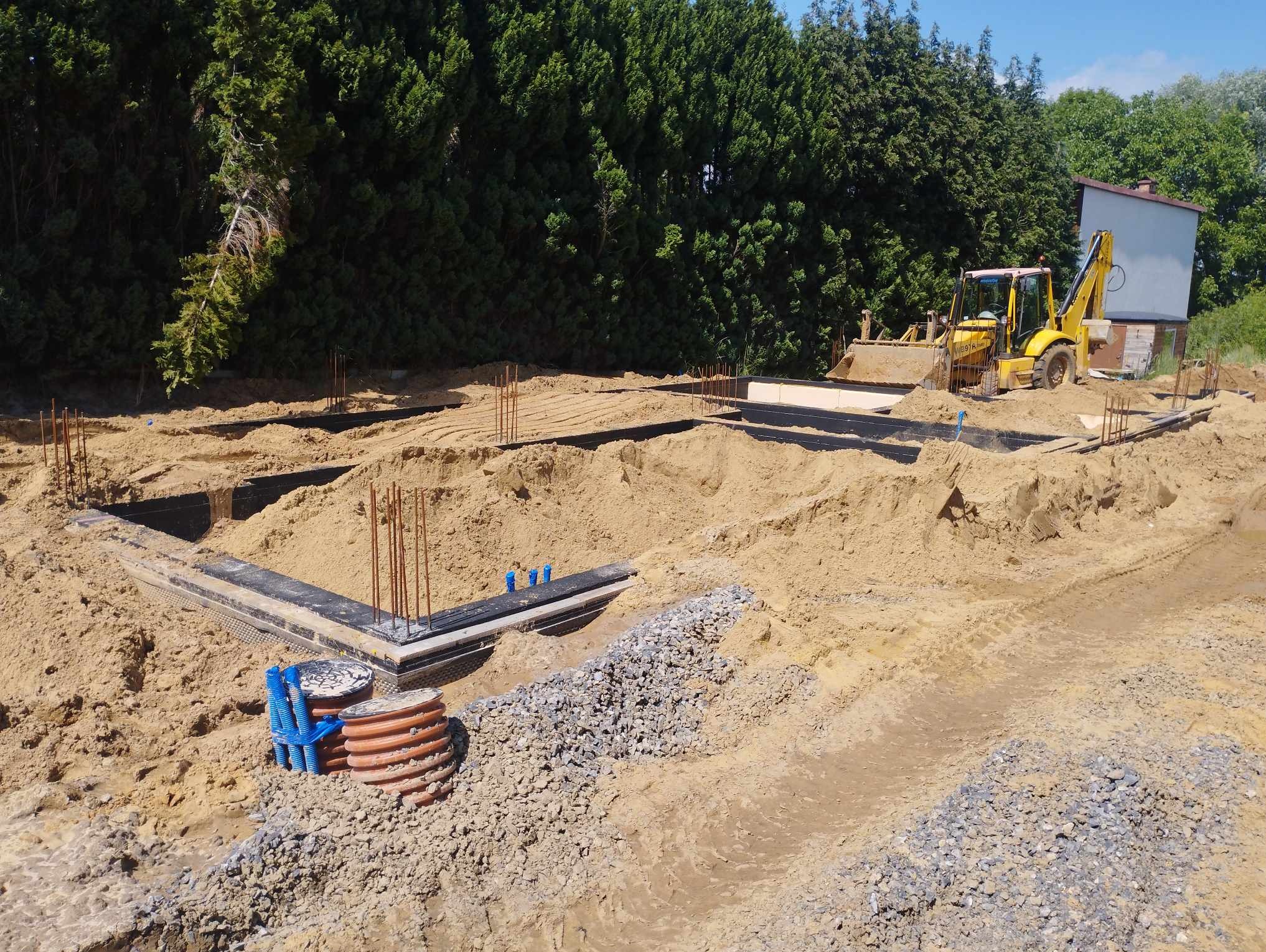 Przygotowanie terenu pod budowę: wykopane fundamenty, szalunki, wystające pręty zbrojeniowe, rury drenażowe oraz koparko-ładowarka w tle.