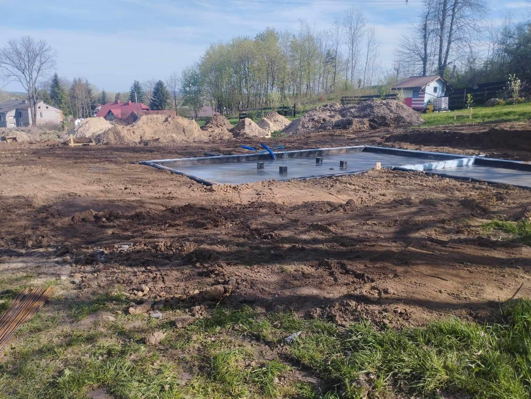 Wylany fundament z widocznymi izolacjami, szalunkiem i wystającymi rurami instalacyjnymi na przygotowanym terenie budowy, w tle pryzmy ziemi i zabudowania.