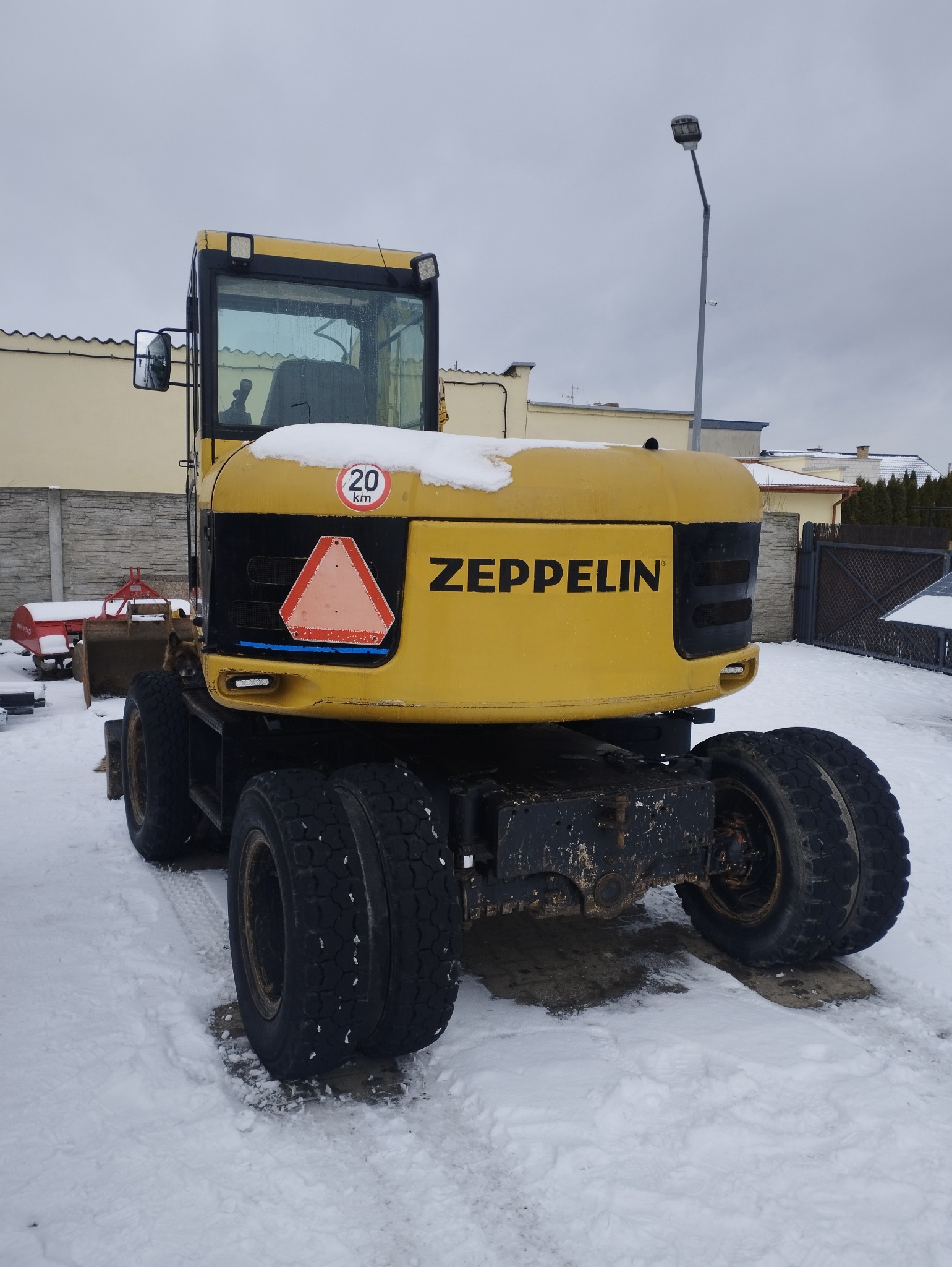 Żółta koparka kołowa marki Zeppelin, widziana z tyłu, stoi na śniegu. Widoczny znak ostrzegawczy i tabliczka z ograniczeniem prędkości 20 km/h.