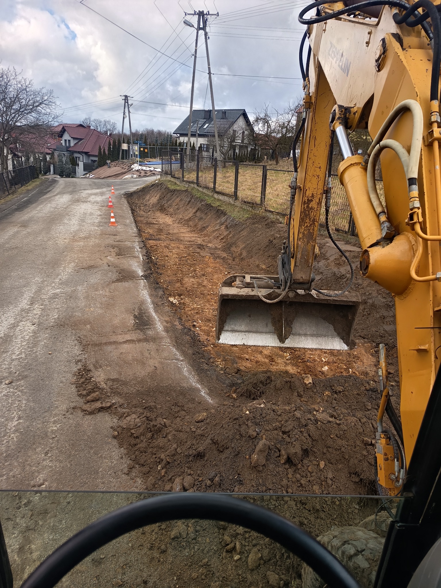 Żółta koparka Zeppelin podczas prac ziemnych przy drodze, widok z kabiny. Wykop wzdłuż drogi, pomarańczowe pachołki, domy w tle.