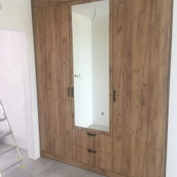 Krzys Meble - Trzydrzwiowa szafa wnękowa z drewnopodobnym dekorem, centralne drzwi z lustrem i dwiema szufladami na dole, widoczna drabina malarska w tle.