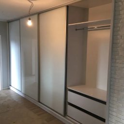 Krzys Meble - Biała szafa przesuwna z lustrzanymi drzwiami, widoczne dwie szuflady i półka z drążkiem na wieszaki, ściana z białej cegły dekoracyjnej po prawej stronie, surowa żarówka zwisająca z sufitu.