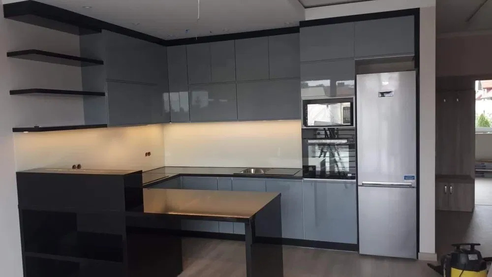 Nowoczesna kuchnia z szarymi, lakierowanymi frontami, czarnym blatem i wyspą, oświetleniem LED podszafkowym i srebrną lodówką w zabudowie, widoczny odkurzacz na podłodze.