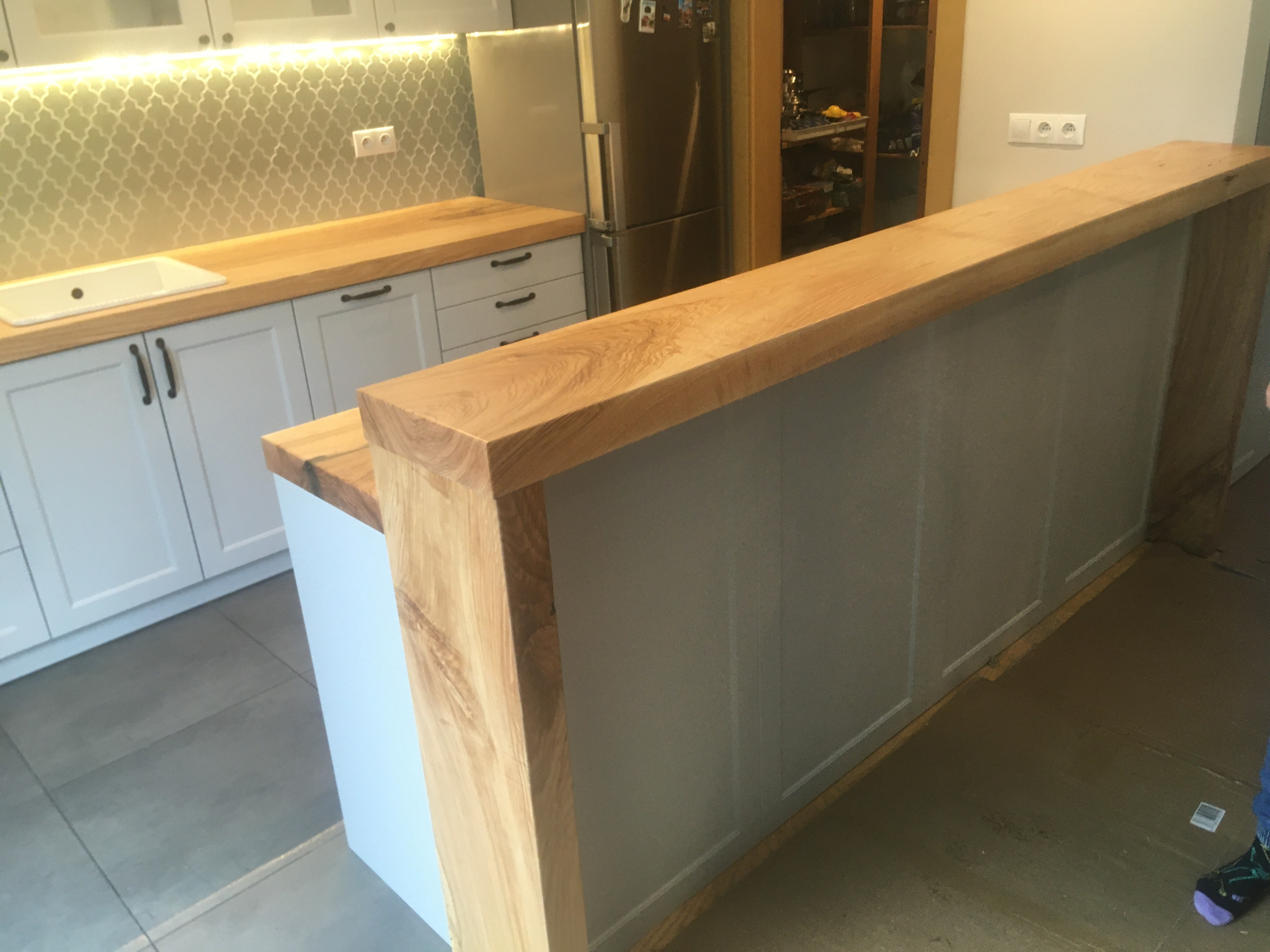 Nowoczesna kuchnia z drewnianym blatem, szarymi szafkami i wyspą z blatem z litego drewna dębowego, widoczne oświetlenie podszafkowe i płytki w marokański wzór.