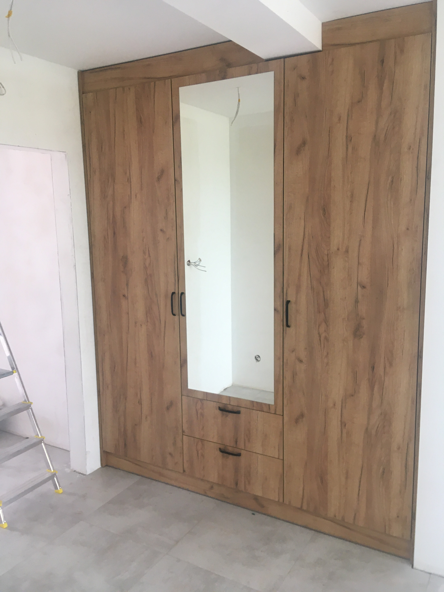 Trzydrzwiowa szafa wnękowa z drewnopodobnym dekorem, centralne drzwi z lustrem i dwiema szufladami na dole, widoczna drabina malarska w tle.