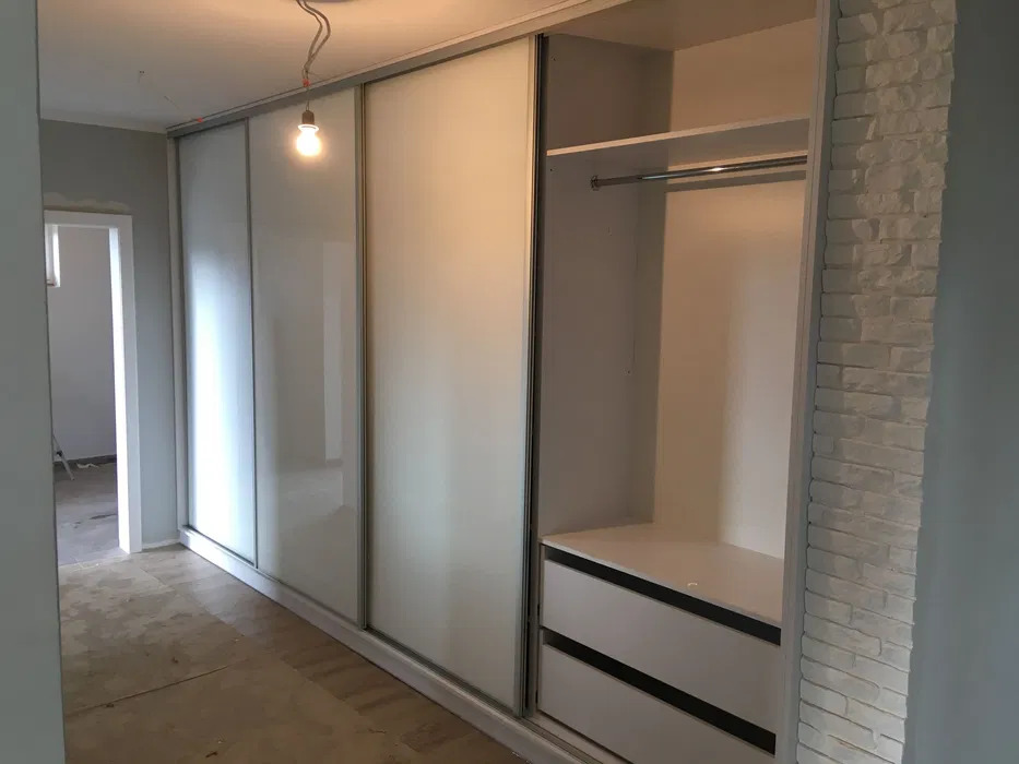 Biała szafa przesuwna z lustrzanymi drzwiami, widoczne dwie szuflady i półka z drążkiem na wieszaki, ściana z białej cegły dekoracyjnej po prawej stronie, surowa żarówka zwisająca z sufitu.