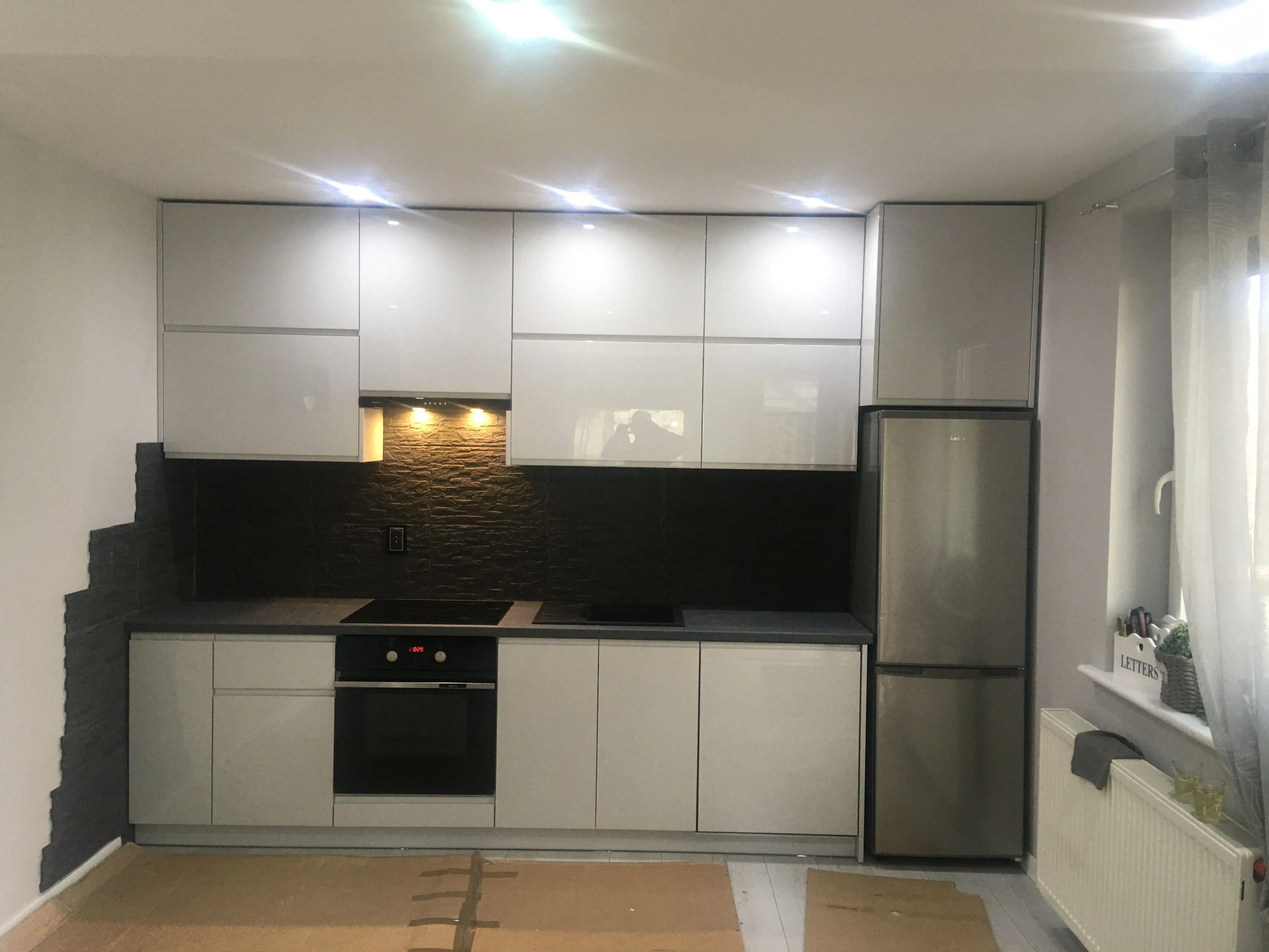 Biała kuchnia z połyskiem, czarny kamienny backsplash, piekarnik z wyświetlaczem 10:29, srebrna lodówka side-by-side, częściowo zakryta podłoga kartonem ochronnym.