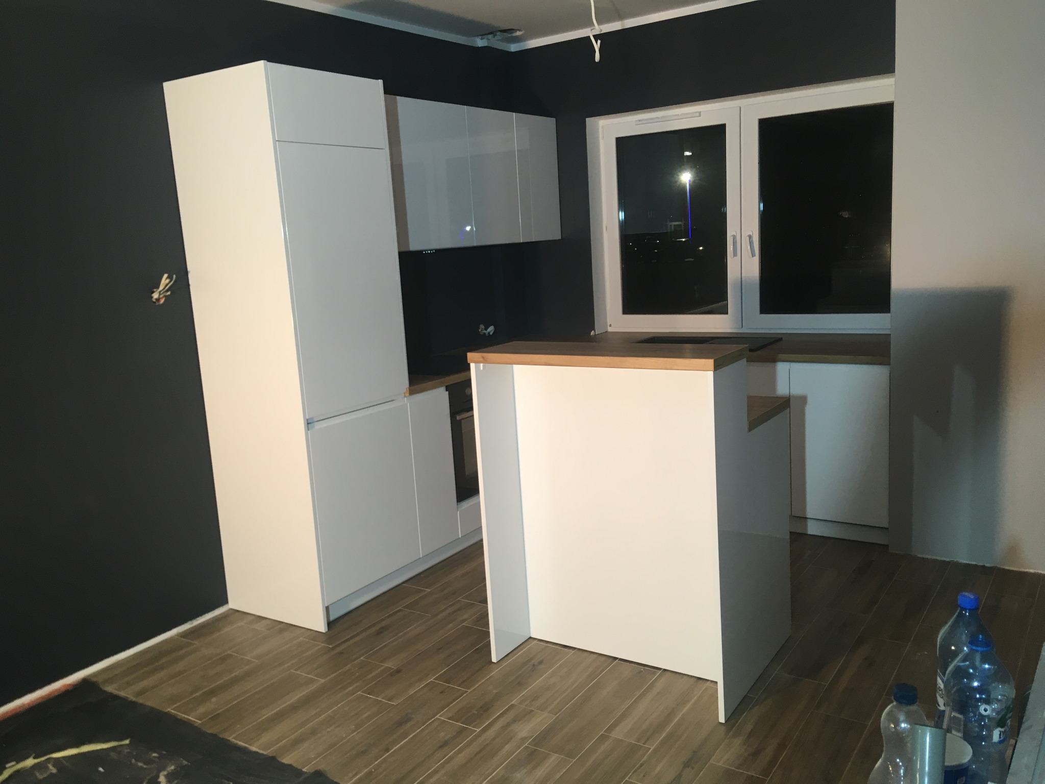 Biała zabudowa kuchenna z wyspą i drewnianym blatem w trakcie montażu, widoczne niedokończone wykończenia i butelki z wodą na podłodze.