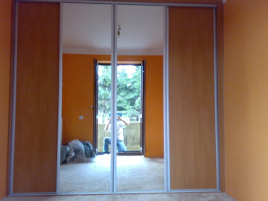 Trzydrzwiowa szafa wnękowa z lustrzanymi drzwiami przesuwnymi, odbijająca pomarańczowe ściany pokoju i osobę robiącą zdjęcie z balkonu.