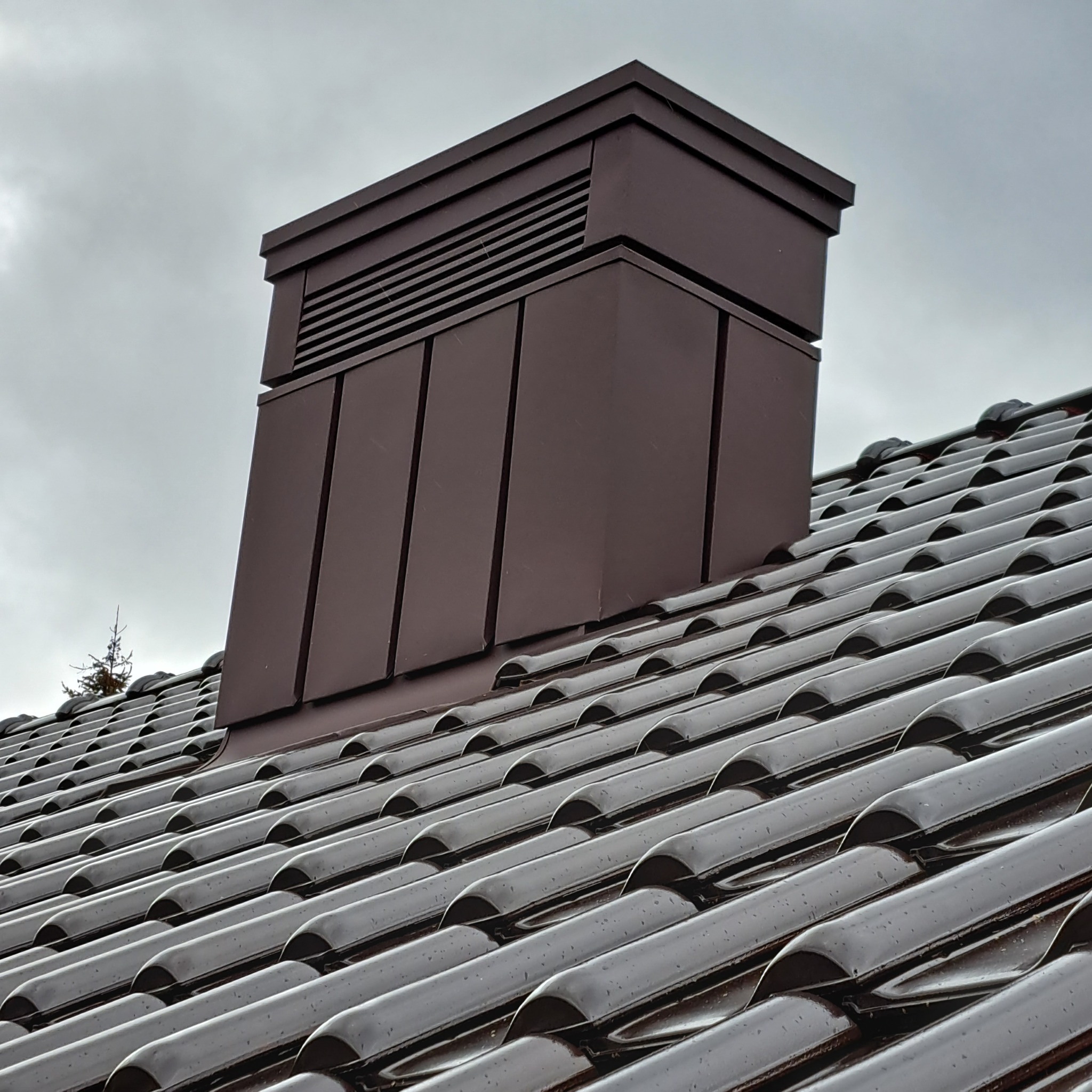 Błyszcząca, brązowa dachówka ceramiczna z nowym, metalowym kominem wentylacyjnym na tle pochmurnego nieba, widoczny fragment drzewka.