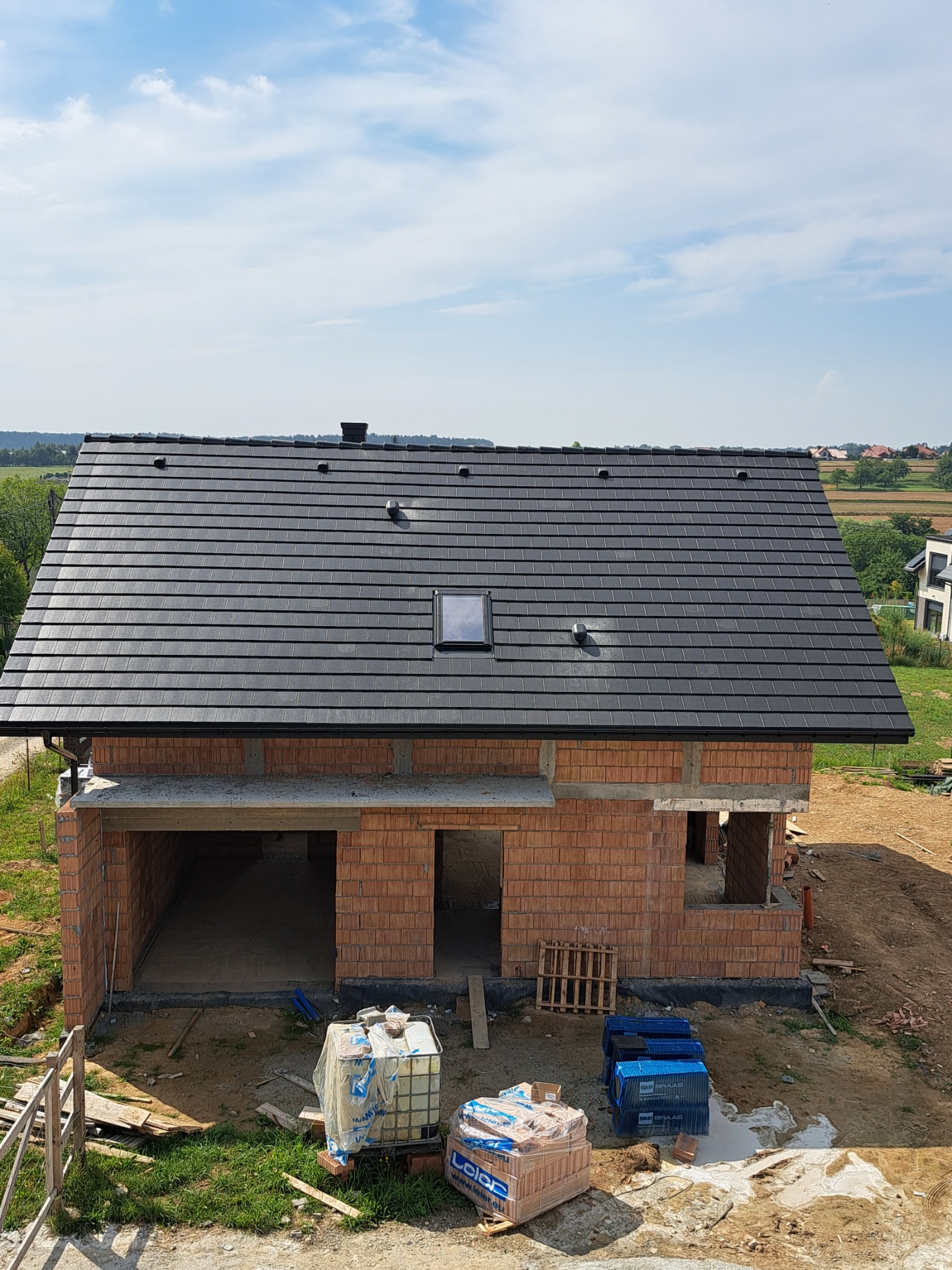 Nowy, czarny dach z dachówki ceramicznej na surowym budynku z czerwonej cegły, widoczne okno dachowe i komin, w tle zielone pola i błękitne niebo.