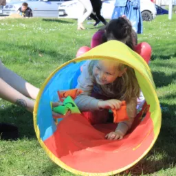Uśmiechnięta blond dziewczynka w tunelu do zabawy na trawie, trzymająca kolorowe woreczki z grochem, z tyłu widoczne inne dzieci i osoba dorosła z tatuażem na przedramieniu.