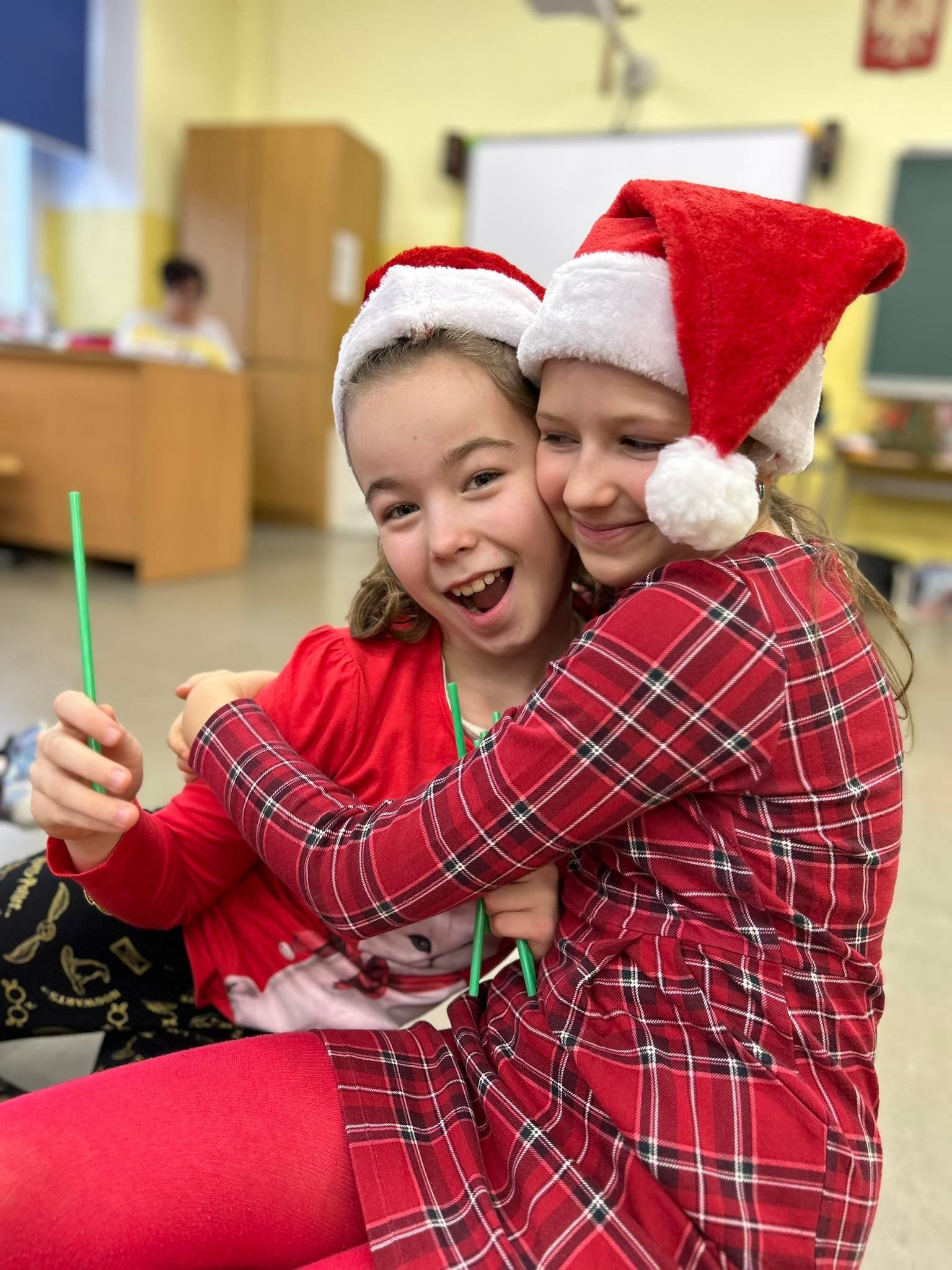 Dwie uśmiechnięte dziewczynki w czerwonych czapkach Mikołaja, obejmujące się w szkolnej klasie, trzymają zielone słomki do napojów.