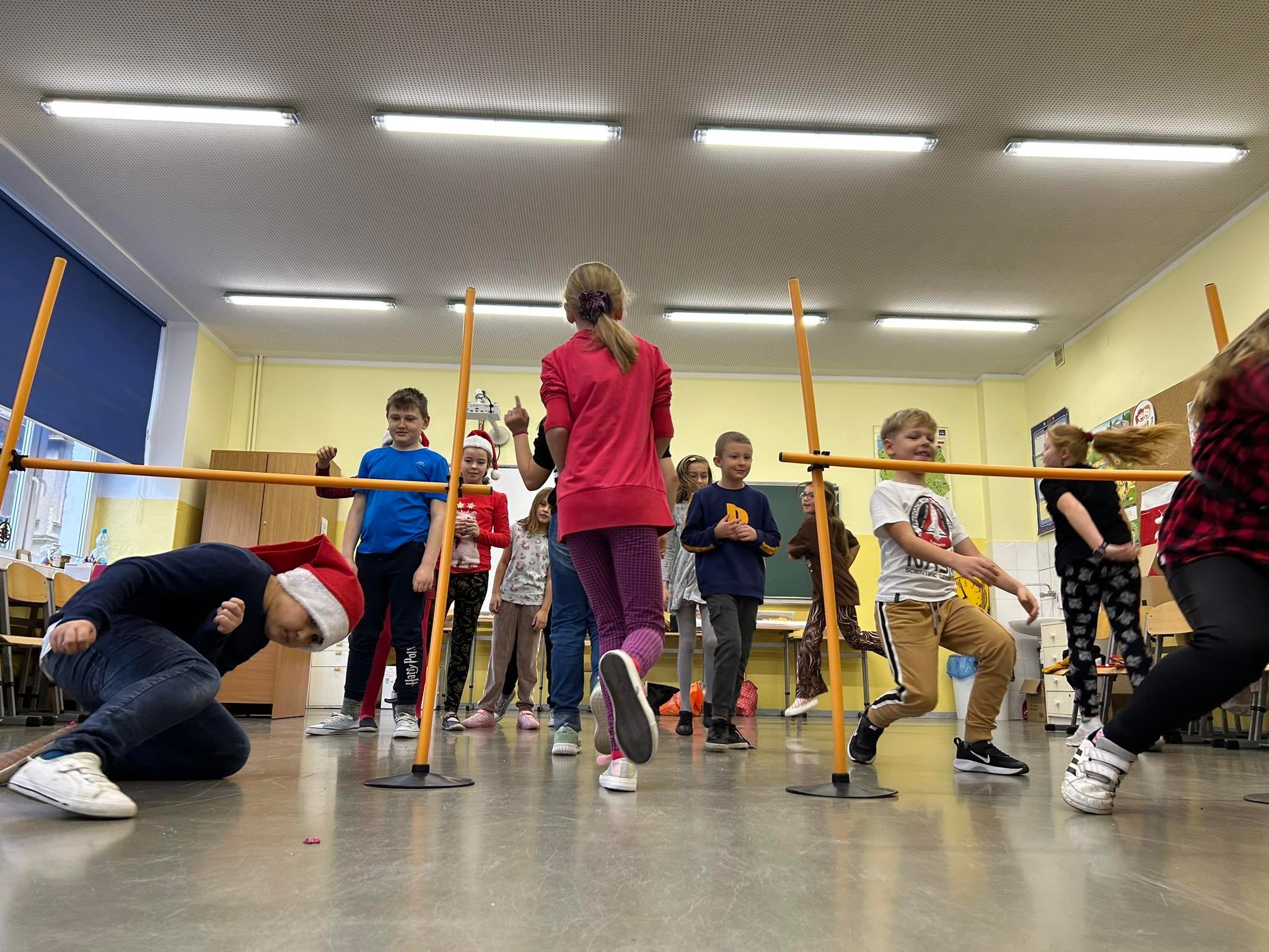 Dzieci biorące udział w zabawie limbo w sali gimnastycznej, jeden chłopiec w czapce Świętego Mikołaja próbuje przejść pod poprzeczką