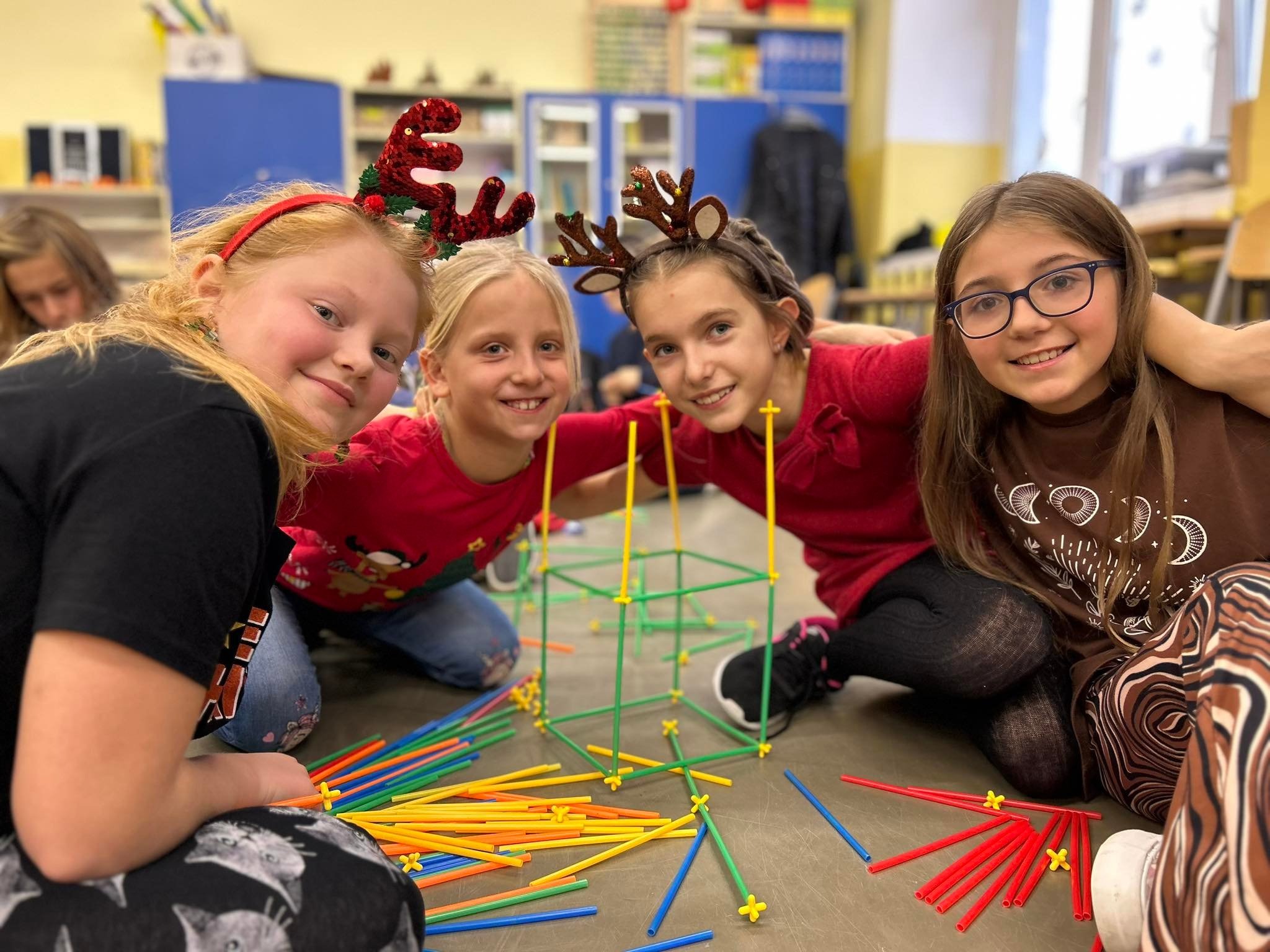 Cztery uśmiechnięte dziewczynki w świątecznych opaskach na głowach siedzą na podłodze i budują konstrukcję z kolorowych patyczków i łączników.