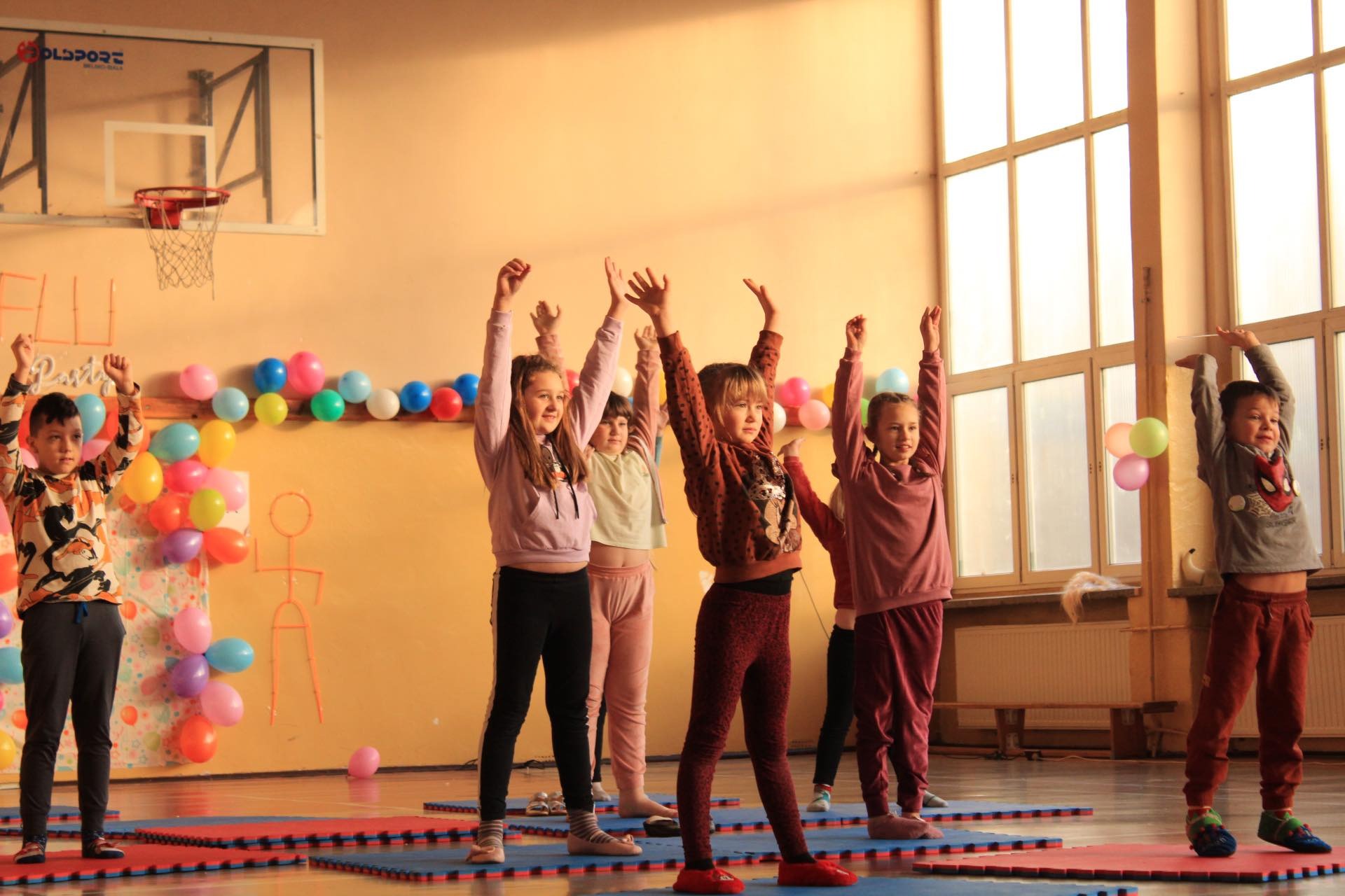 Grupa dzieci w różnym wieku ćwiczy w sali gimnastycznej, unosząc ręce do góry na tle dekoracji z balonów.