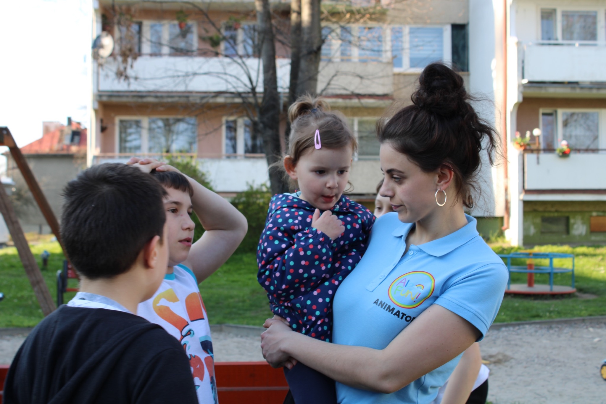Kobieta w błękitnej koszulce z logo firmy trzyma na rękach małą dziewczynkę w kurtce w kropki, wokół stoją dzieci na placu zabaw.