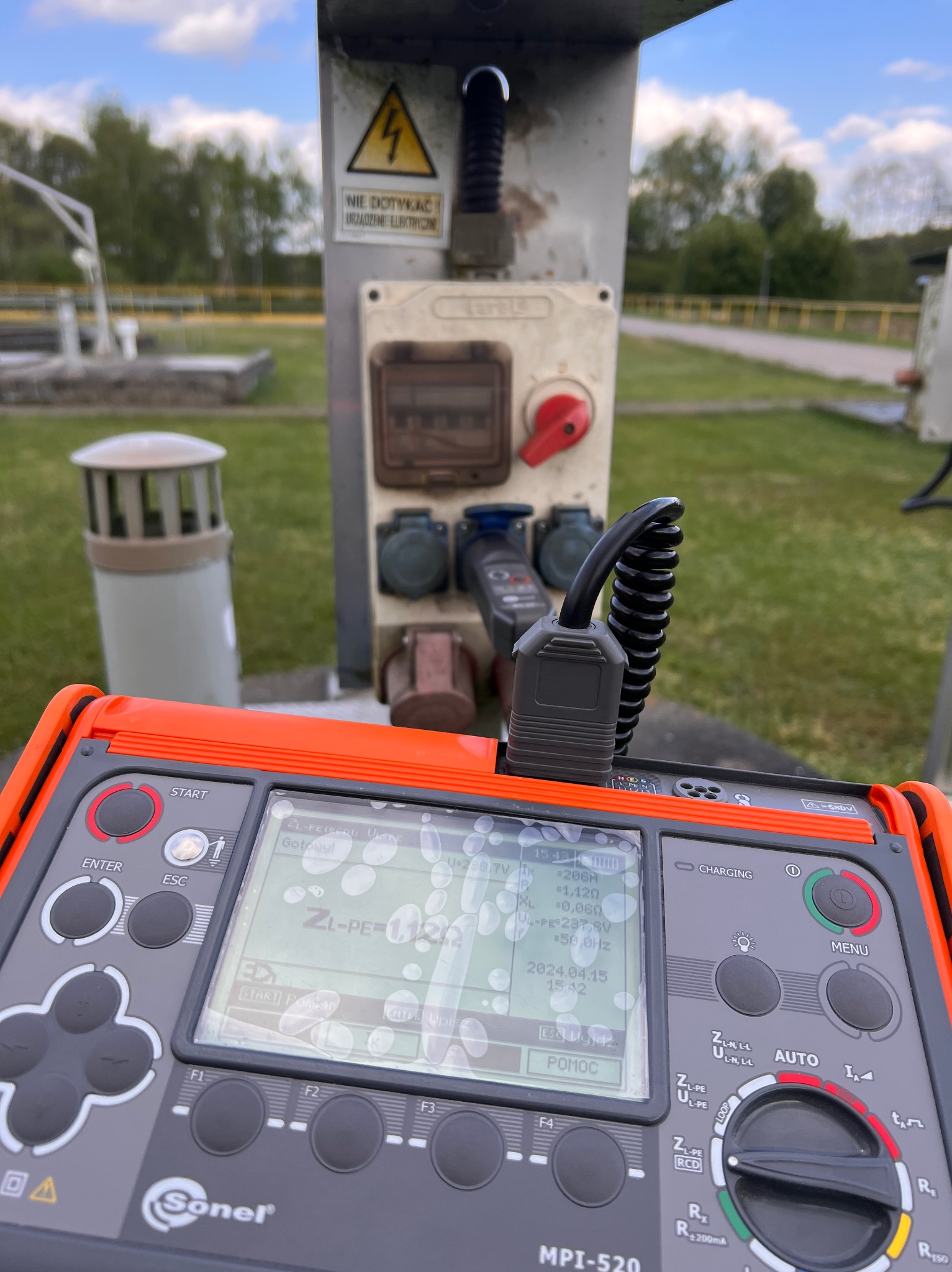 Miernik Sonel MPI-520 na tle skrzynki elektrycznej z gniazdami i ostrzeżeniem o napięciu, widoczne wyniki pomiarów na wyświetlaczu.