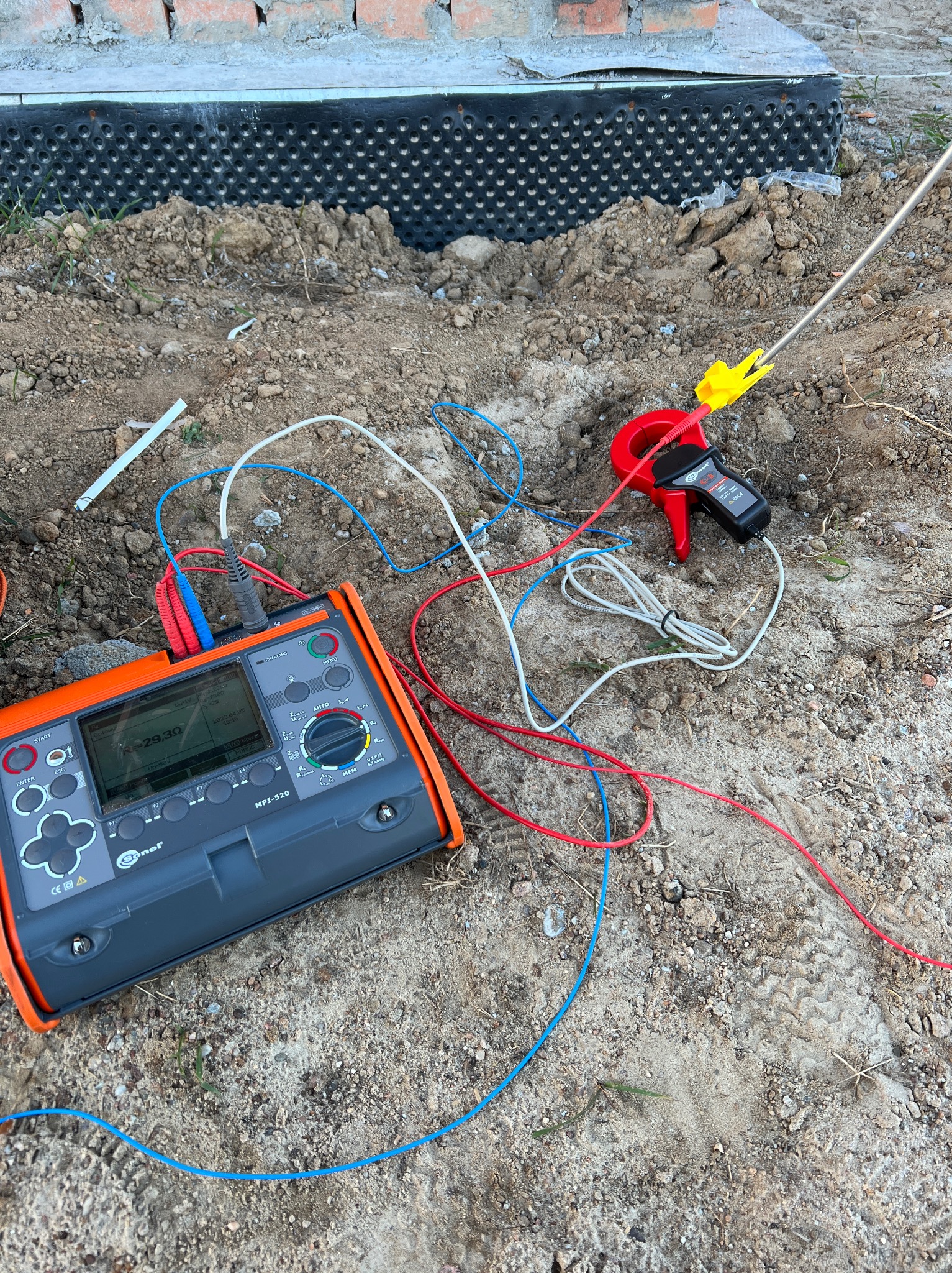 Miernik wielofunkcyjny Sonel MPI-520 z podłączonymi przewodami pomiarowymi i cęgami prądowymi na ziemi, w tle fundament budynku z izolacją pionową.