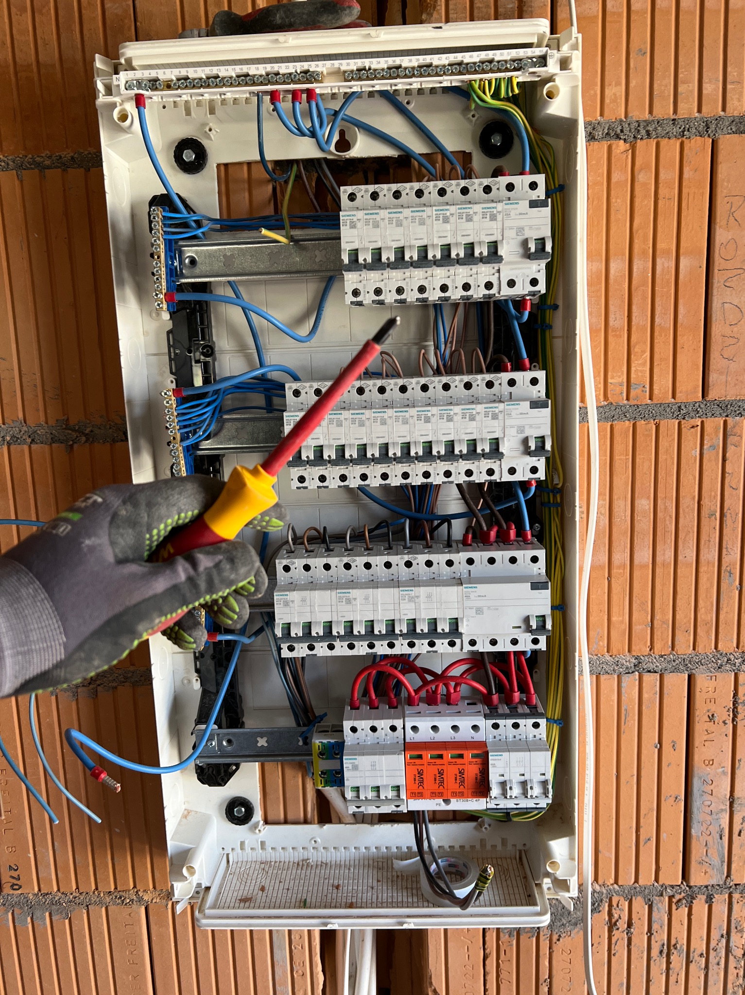 Otwarta skrzynka rozdzielcza z bezpiecznikami i przewodami, ręka w roboczej rękawicy trzyma śrubokręt z izolowanym grotem, mur z cegły w tle.