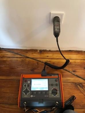 Miernik elektryczny z pomarańczową obudową podłączony do gniazdka elektrycznego na tle drewnianej ściany.