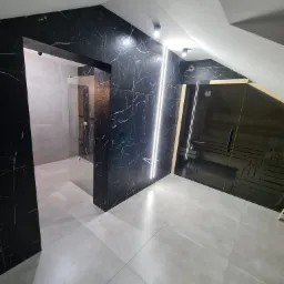 Nowoczesna łazienka w odcieniach szarości i czerni z marmurowymi płytkami, szklanym prysznicem i sauną w skosie poddasza, oświetlona punktowymi lampami i pionowymi paskami LED.
