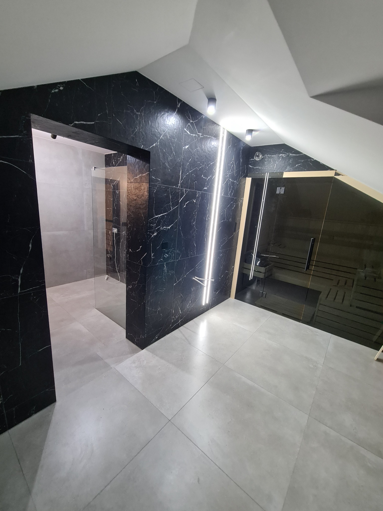 Nowoczesna łazienka w odcieniach szarości i czerni z marmurowymi płytkami, szklanym prysznicem i sauną w skosie poddasza, oświetlona punktowymi lampami i pionowymi paskami LED.