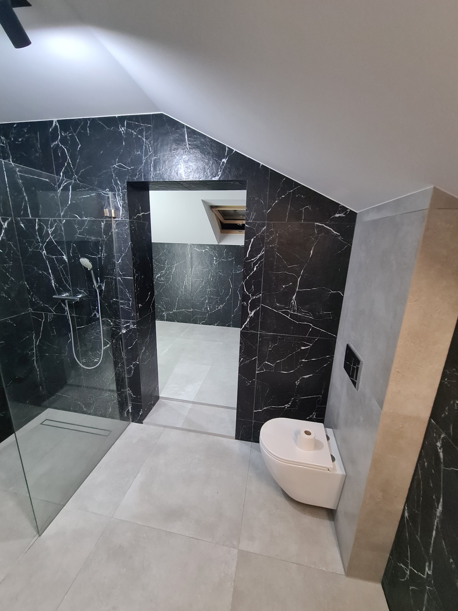 Nowoczesna łazienka na poddaszu z czarnymi płytkami imitującymi marmur i kabiną prysznicową typu walk-in, wisząca toaleta z rolką papieru, szara podłoga i ściany w neutralnych odcieniach...