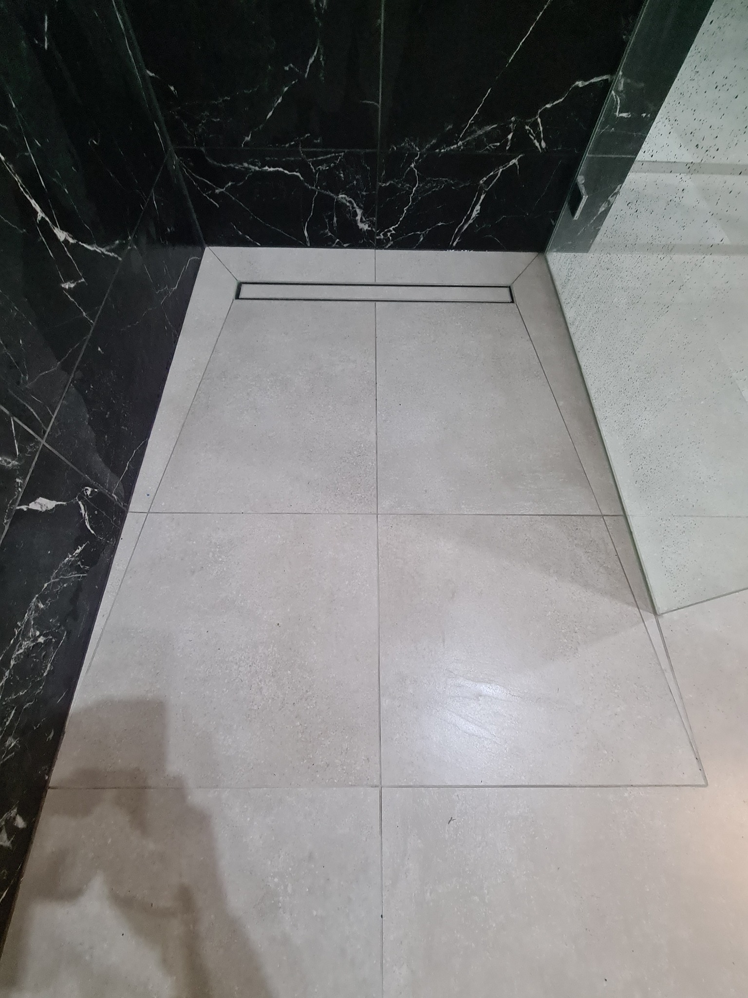 Wykończona kabina prysznicowa walk-in z czarnymi marmurowymi płytkami na ścianach i jasnymi, kwadratowymi płytkami podłogowymi oraz liniowym odpływem.