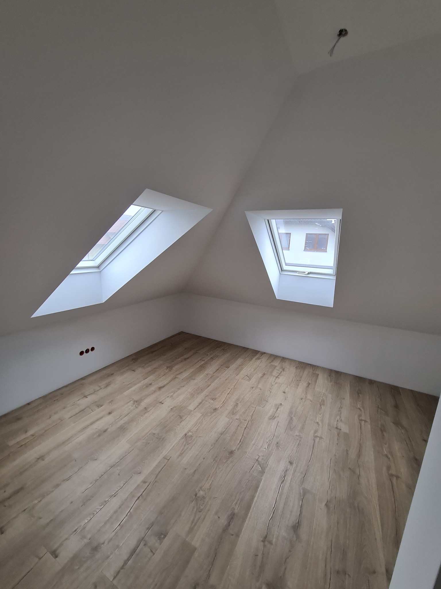 Poddasze z dwoma oknami dachowymi i wykończoną podłogą panelową w odcieniach brązu, świeżo pomalowane ściany na biało z widocznymi otworami na gniazdka elektryczne.