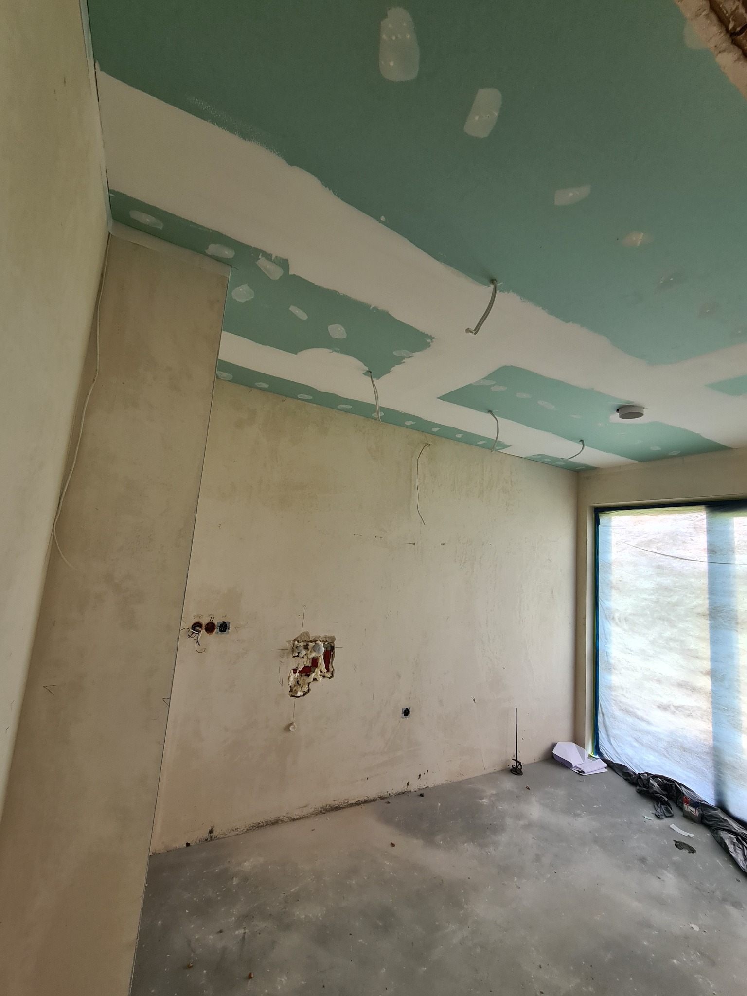 Wnętrze w trakcie prac wykończeniowych: surowa ściana, częściowo zamontowane płyty na suficie, wystające kable elektryczne, niezasłonięte otwory na gniazdka, okno zasłonięte folią ochronną, podłoga...