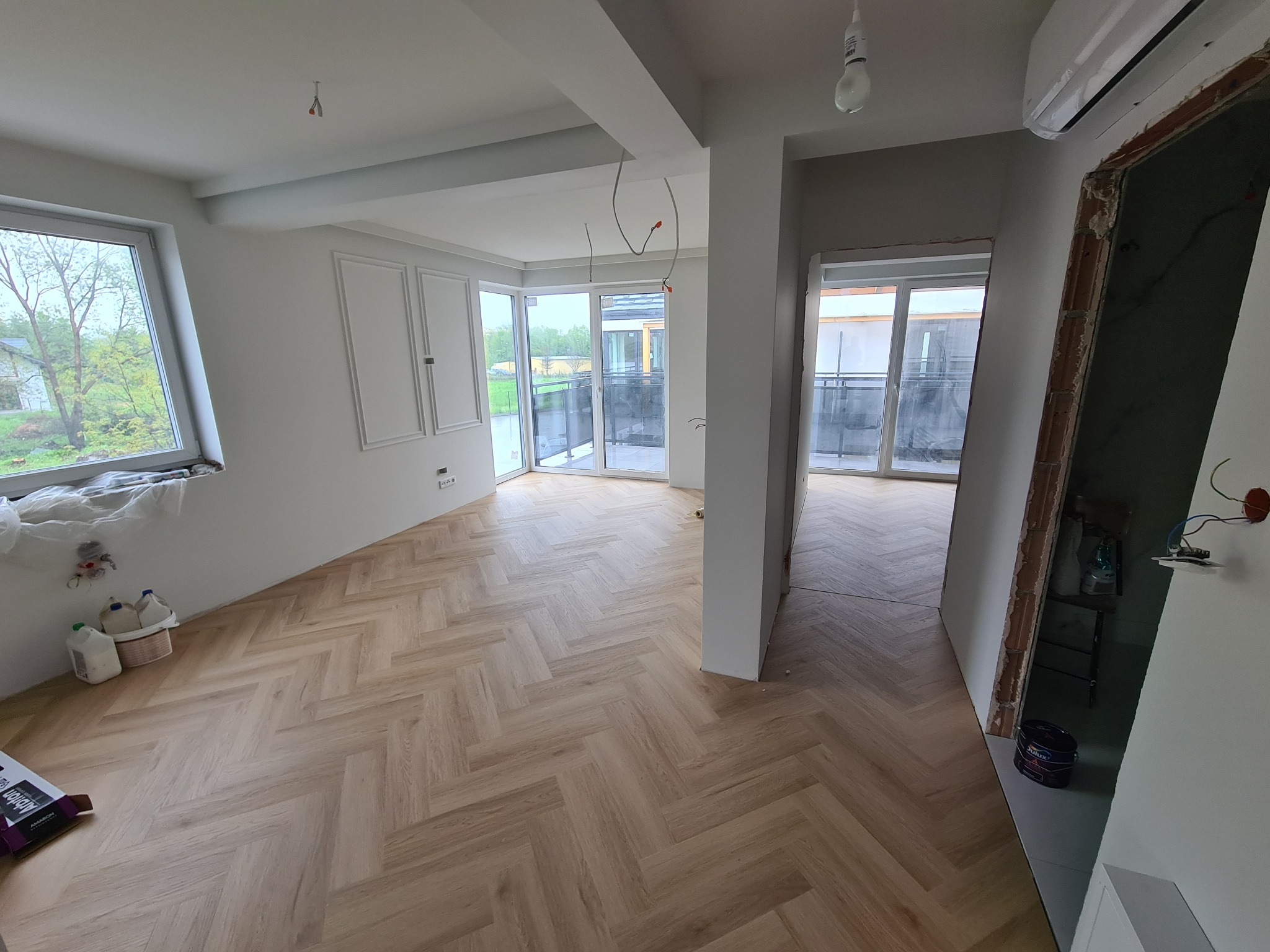 Jasne panele ułożone w jodełkę w przestronnym, częściowo wykończonym wnętrzu z białymi ścianami i dużymi oknami; widoczne elementy instalacji elektrycznej i puszki z farbą.