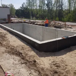 Wykop z betonowymi ścianami fundamentowymi na piaszczystym gruncie w Mikstacie, widoczny pracownik w pomarańczowej kamizelce i drzewa w tle.