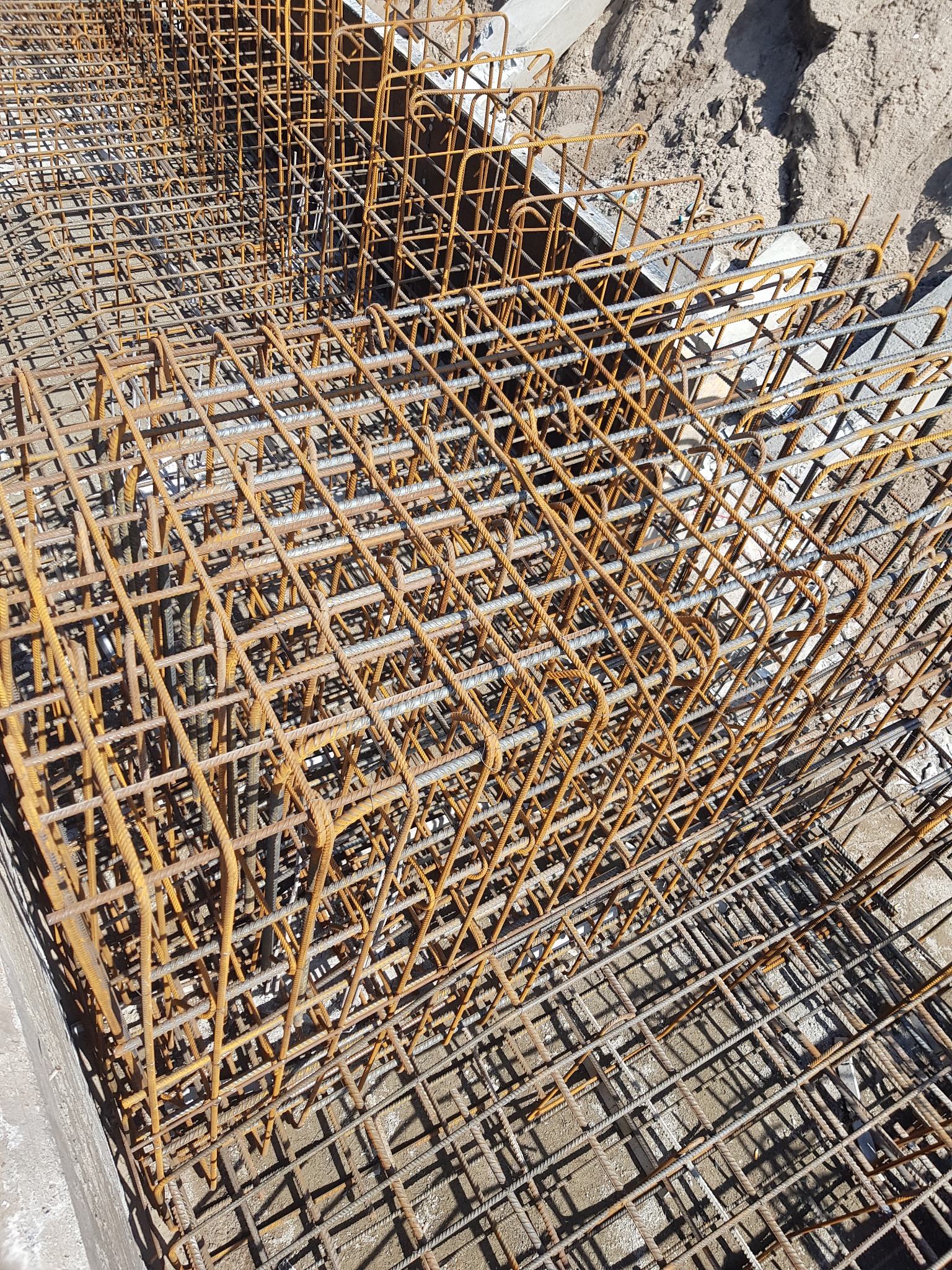 Siatka zbrojeniowa ułożona w szalunku fundamentowym, widoczne zardzewienia na prętach i piasek w tle.