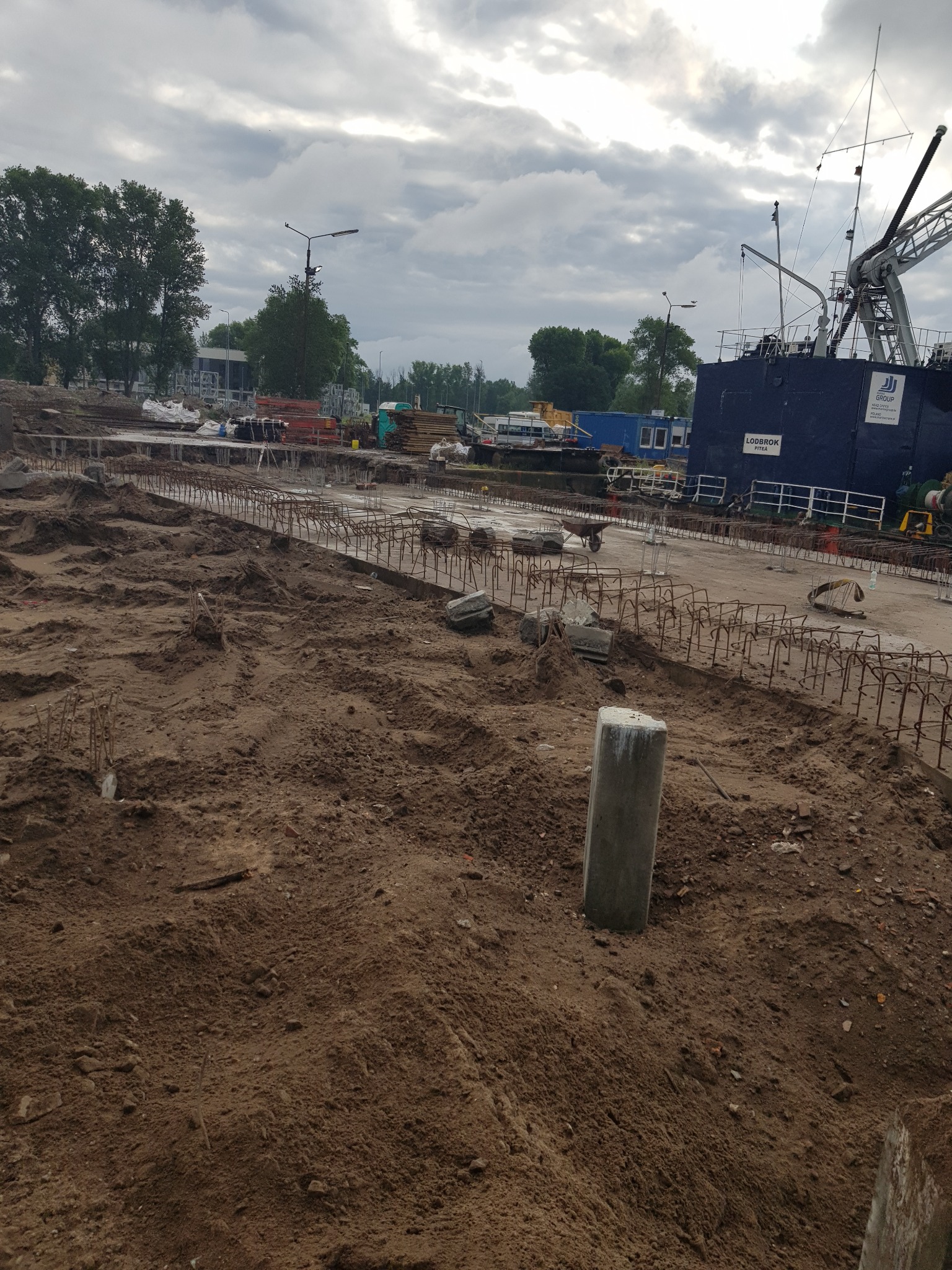 Przygotowanie terenu pod budowę: widoczne zbrojenia, fundamenty i ciężki sprzęt w tle, pochmurne niebo.