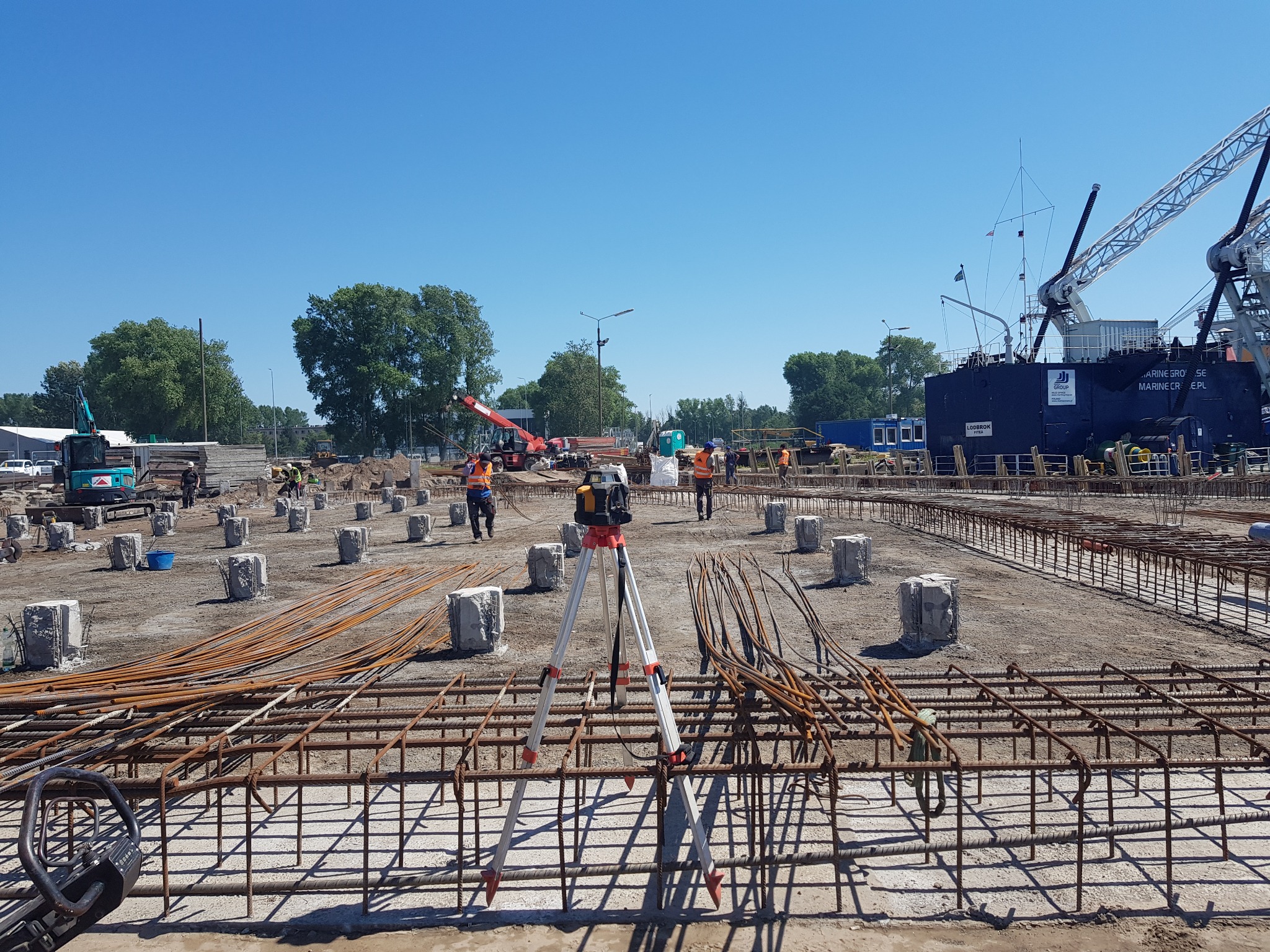 Rozległy plac budowy z widoczną siatką zbrojeniową, betonowymi podporami, pracownikami w odblaskowych kamizelkach oraz geodezyjnym niwelatorem na statywie na pierwszym planie.